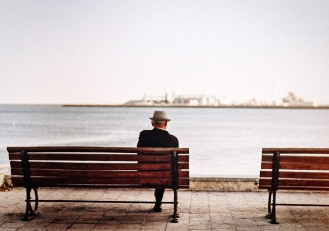 August-29-2019-Elder-Trust elderly man sitting on a bench overlooking a city harbor