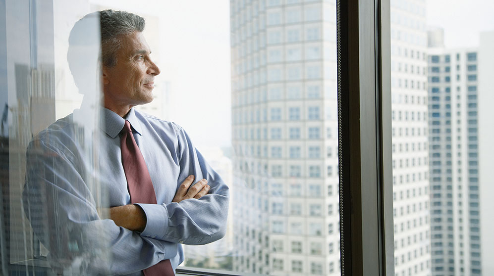 Business man with arms crossed across chest looking out at city buildings.