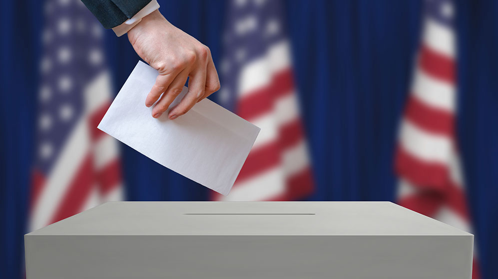 Hand dropping ballot envelope in a box with American flags in the background.