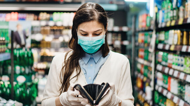 Woman wearing surgical gloves and mask in grocery aisle checking wallet.