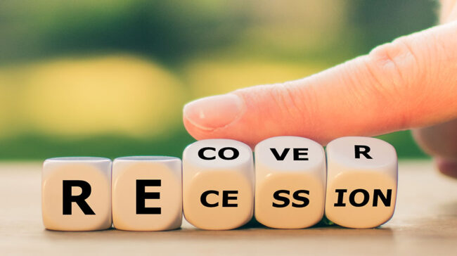 Finger turning over game die that could spell R-E-CO-VE-R or R-E-CE-SS-ION.