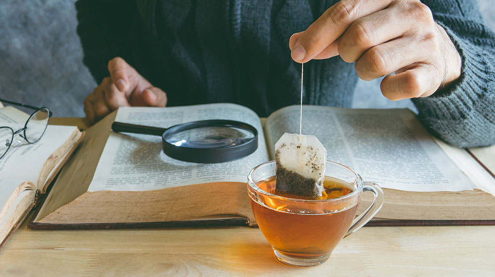 Hand dunking a tea bag in a glass tea cup with open book with small type needing a magnifying glass to read.