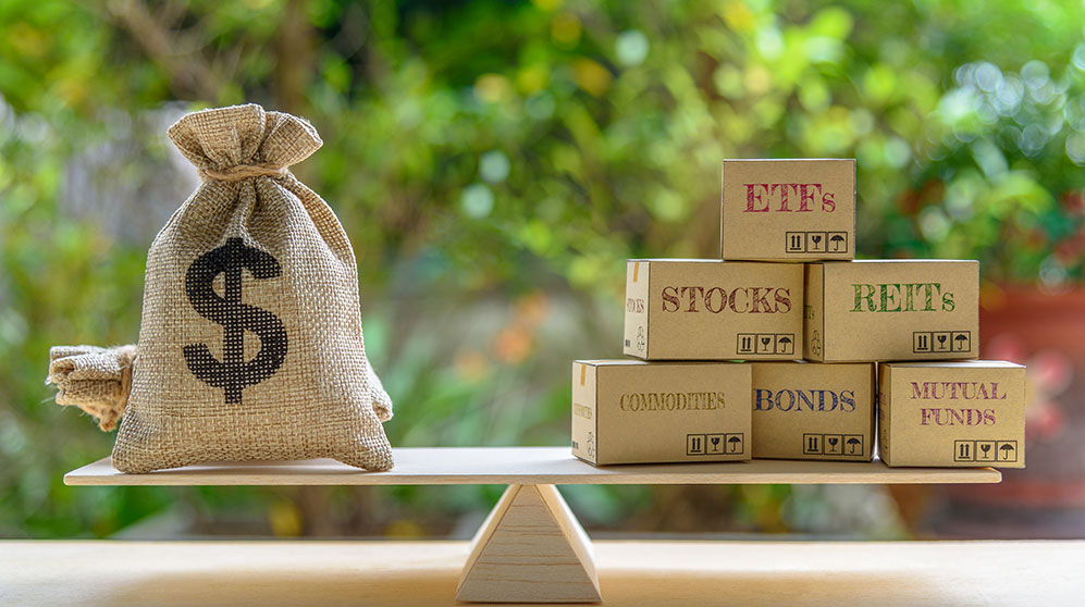 Evenly balanced board over a pointed fulcrum, a burlap bag with $ label on one side, and boxes on other side with labels: ETFs, Stocks, REITs, Commodities, Bonds, Mutual Funds.