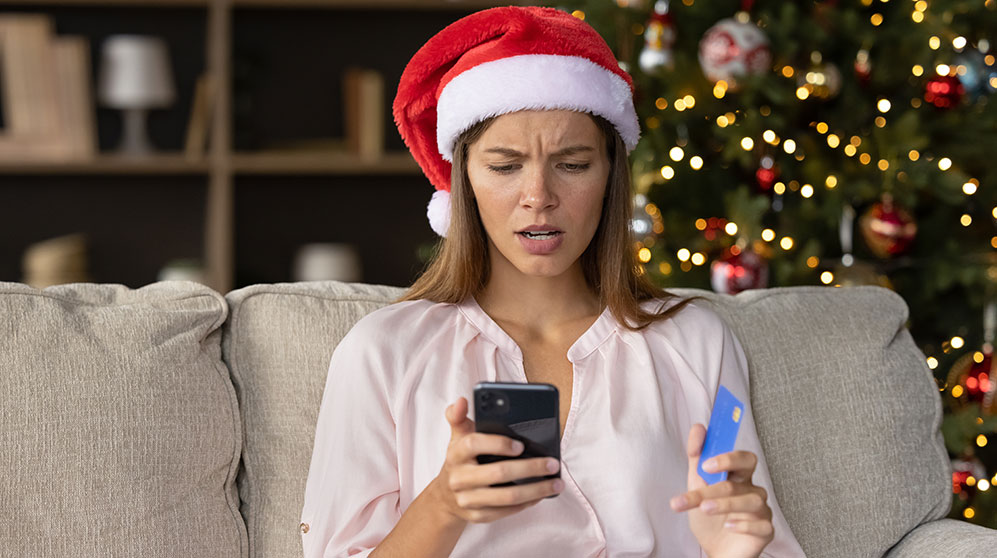 Woman looking confused while seated on couch wearing a santa hat on making an online purchase