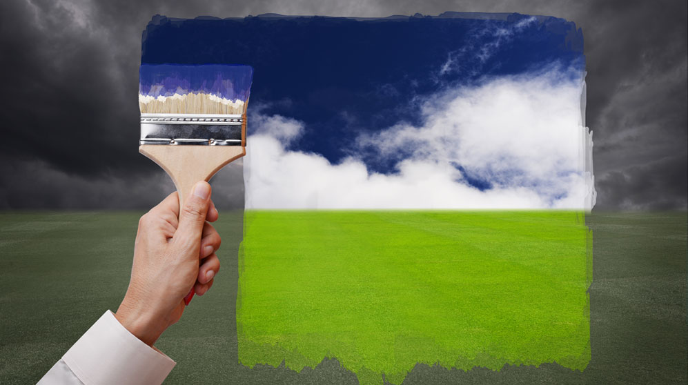 hand with paintbrush painting a bright blue sky and green grass on a drab horizon