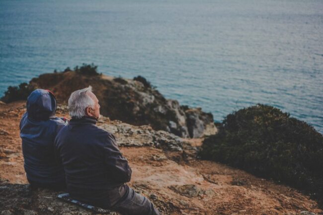Elder couple sitting on cliff looking out at sea.