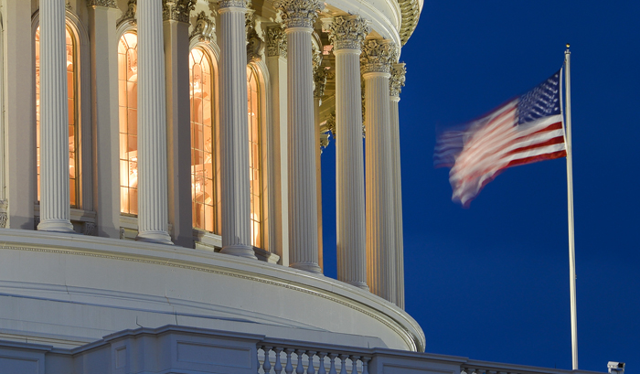 Partial facade of White House lit from within at night with blowing American flag.